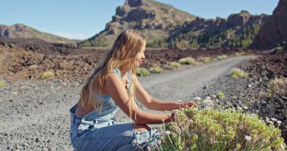 A young woman picks and smell a flower then walks down dirt road near large boulders