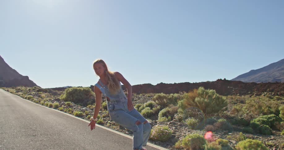 Blonde woman with strappy denim jumpsuit rides on skateboard on asphalt road