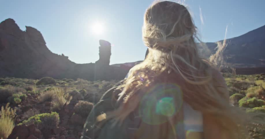 A blond woman wearing a backpack is walking among rock formations and mountains with the sun shining brightly upon her