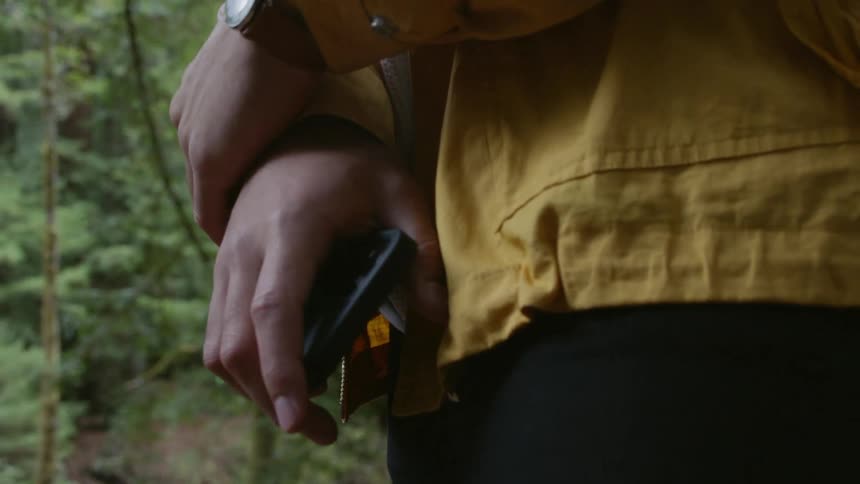 A man holds his cell phone and crosses his arms while standing in a forest