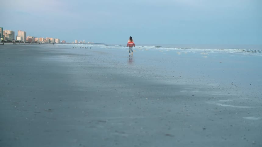A person walking on a beach with the ocean buildings the sky and clouds in the background