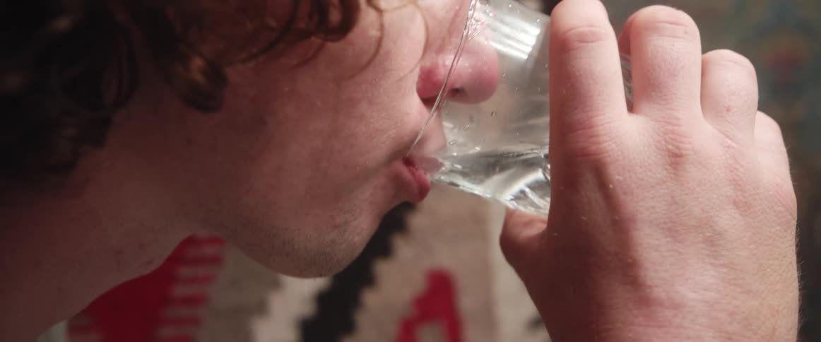 A man in a room drinking a glass of water with a wall in the background