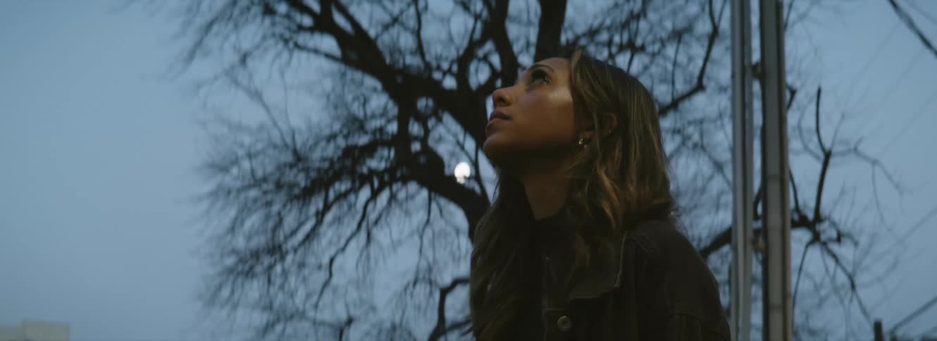 A woman looking up with trees buildings the moon the sky and clouds in the background