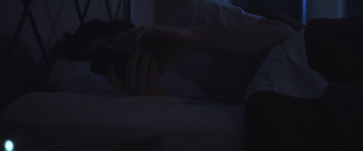 A man in a room laying in bed looking at a smart phone with a wall in the background
