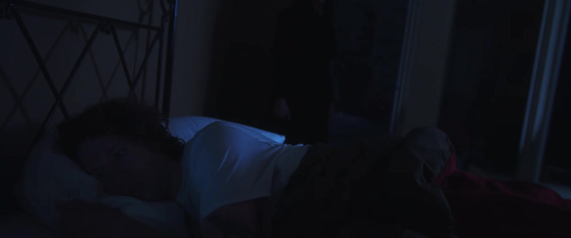 A man in a room laying in a bed with a woman walking and a wall in the background