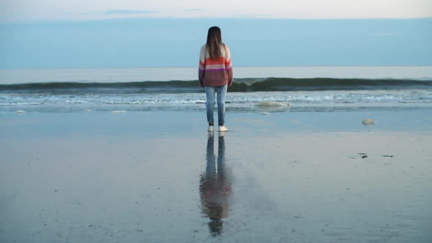 Young woman wearing colorful sweater standing on the coast of a beach near the ocean at sunset