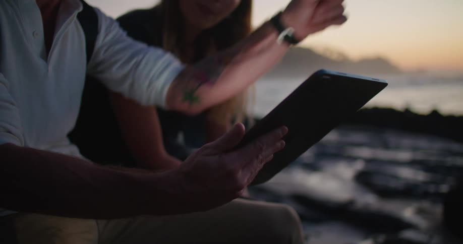 Young couple using a tablet sitting on the rocks near the coast in front of the ocean at sunset