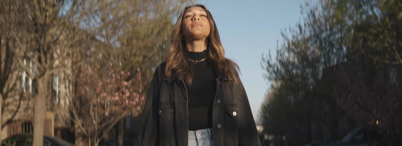 Woman with wavy hair black sweater and denim coat lifts her head up eyes closed in the city street