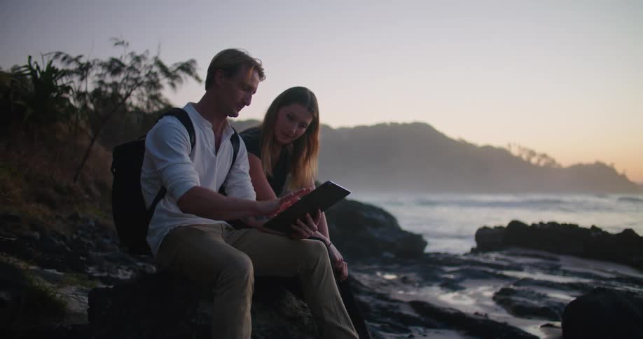 Couple sitting on the rocks near the beach at sunset using a tablet