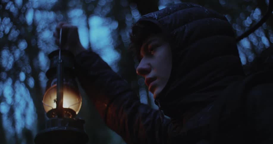 Man wears coat and hood standing in forest holding lantern under rain