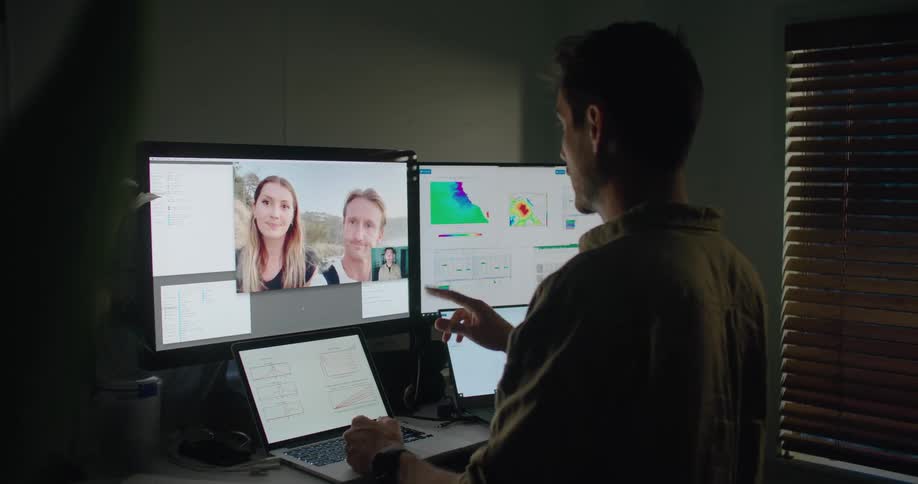 Man sitting at the desk in the office in front of multiple screens on a video call with coworkers