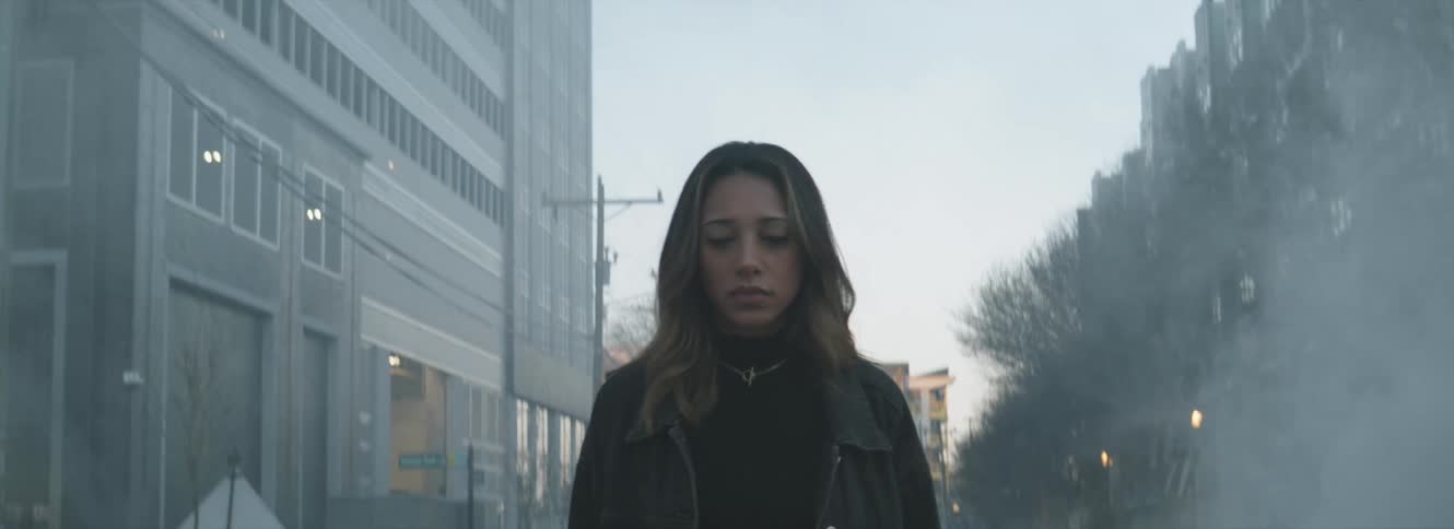 A woman walking on a city street with smoke with buildings trees the sky and clouds in the background