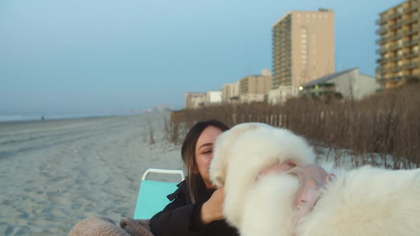 Young woman sitting on a chair in the beach cuddles a white fluffy dog