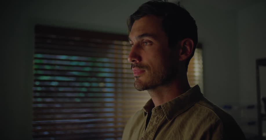 Man with stubble beard and khaki shirt makes video call with man and woman on desktop in dim lit room
