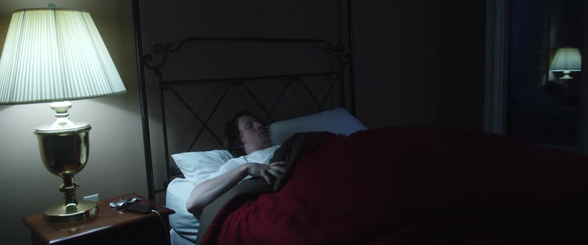 A man in a room laying on a bed and taking off a lamp with a wall and window in the background
