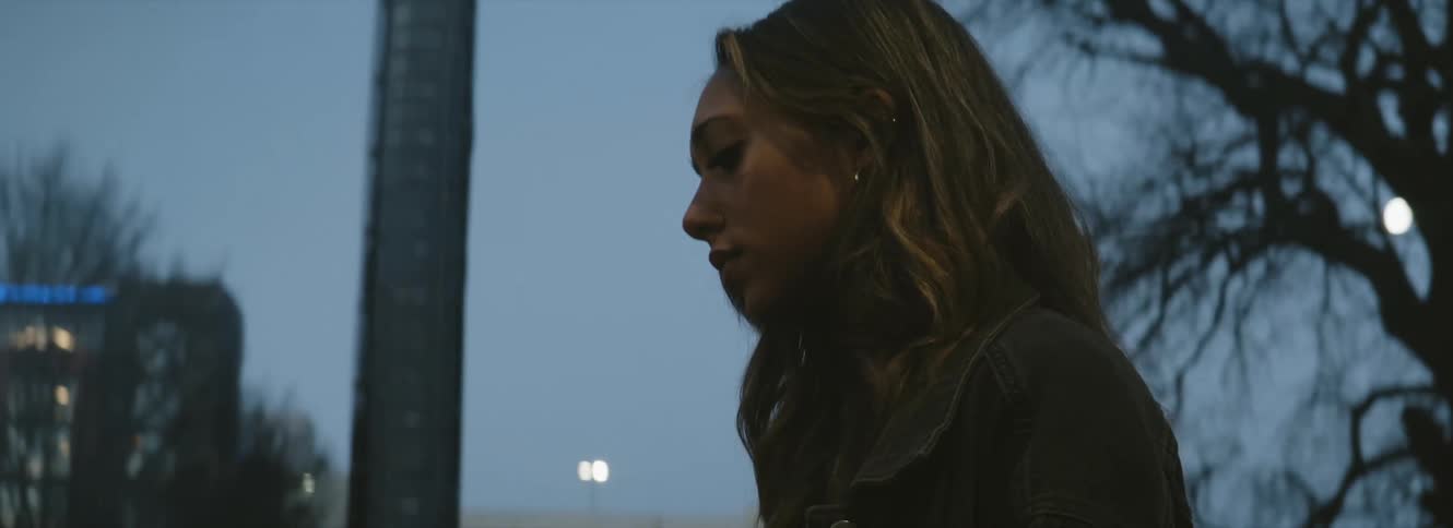 A sad woman stands outdoors in a city on a cloudy evening