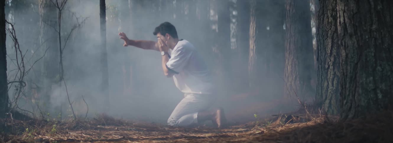 Man wearing white clothes kneeling on the forest floor surrounded by smoke