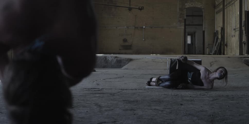 A man in a room with his hands tied with rope and hanging upside down with a woman tied to a chair on the floor with a wall and doorway in the background