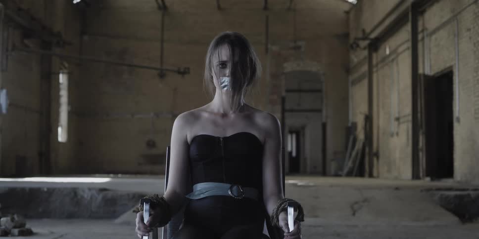Scared woman tied to a chair inside an abandoned warehouse tries to scream
