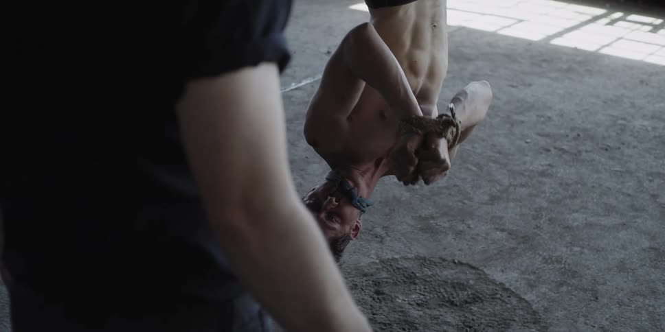 Person walks towards a scared shirtless man tied up hanging upside down inside a warehouse