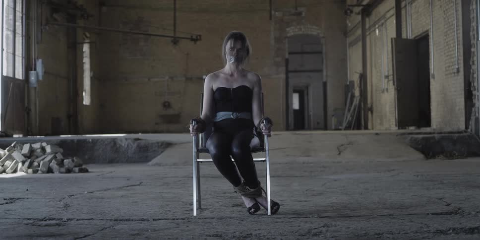 A woman with her mouth taped shut and her ankles tied together sits tied to a chair in an empty building