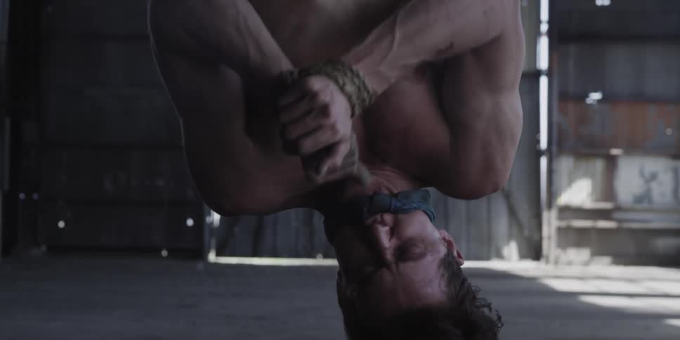 A man bound and gagged is hanging upside down in a barn struggling to cut the rope with a glass shard