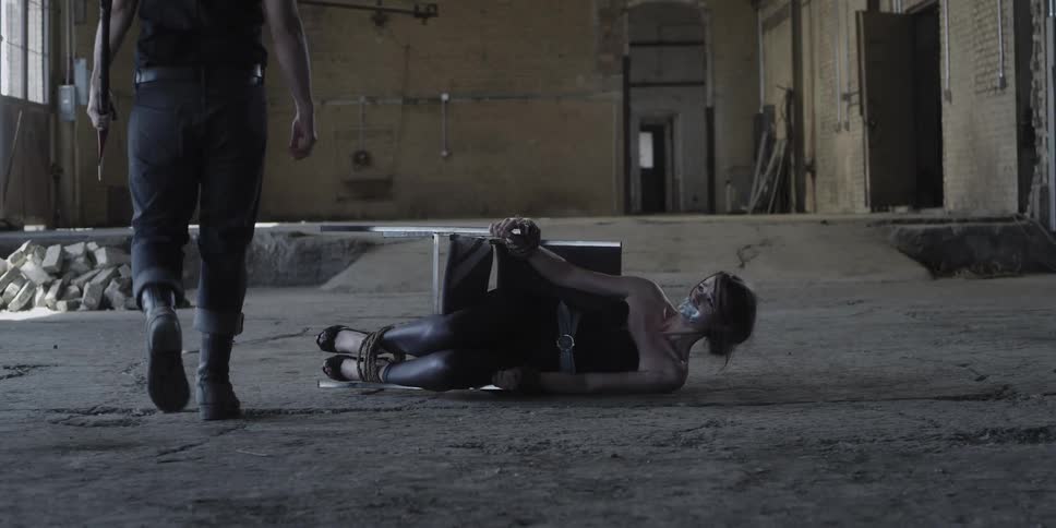 Man carrying an axe walks towards a woman laying on the floor tied to a chair inside a warehouse