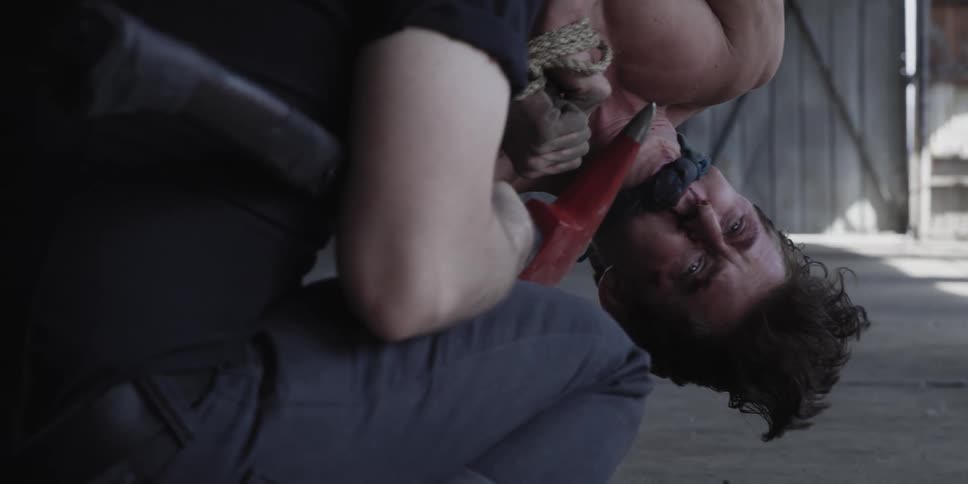 Scared shirtless man gagged and tied up hanging upside down near a person with an ax