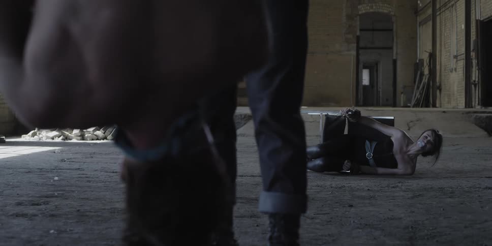 A man tied up upside down and a woman tied to a chair on the ground with a man with an ax between them