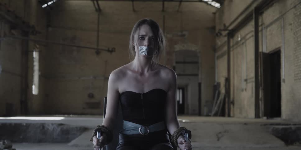 Scared young blonde woman gagged and tied to a chair inside a warehouse
