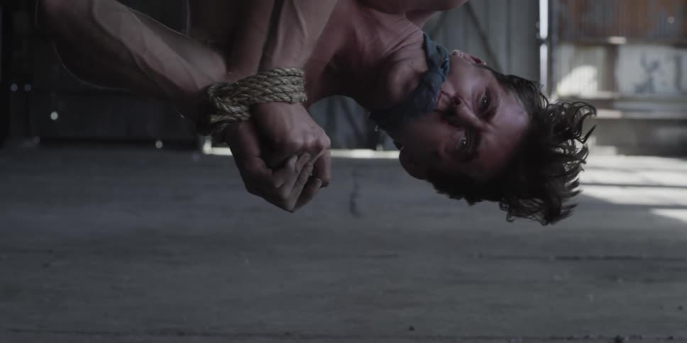 A man in a room hanging upside down with his hands tied up with rope with a wall and door in the background