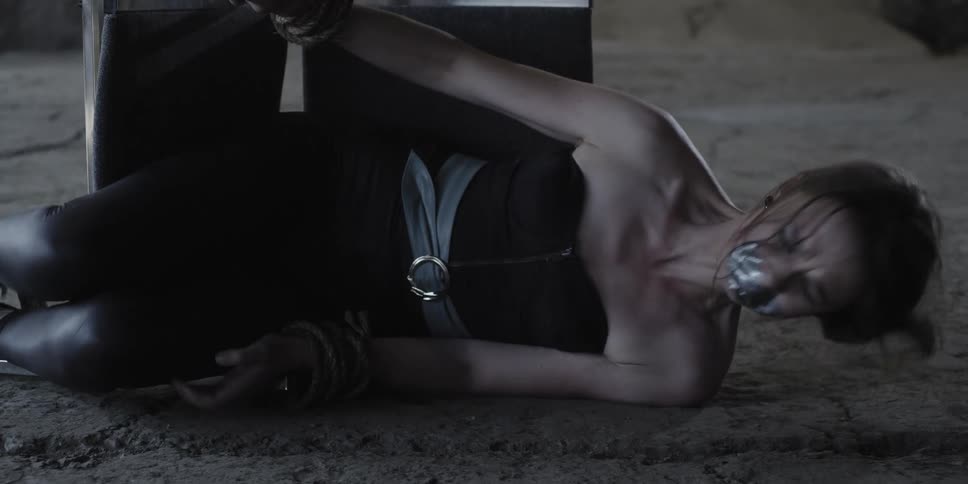 A woman in a room on the floor with her mouth duct taped with a wall in the background