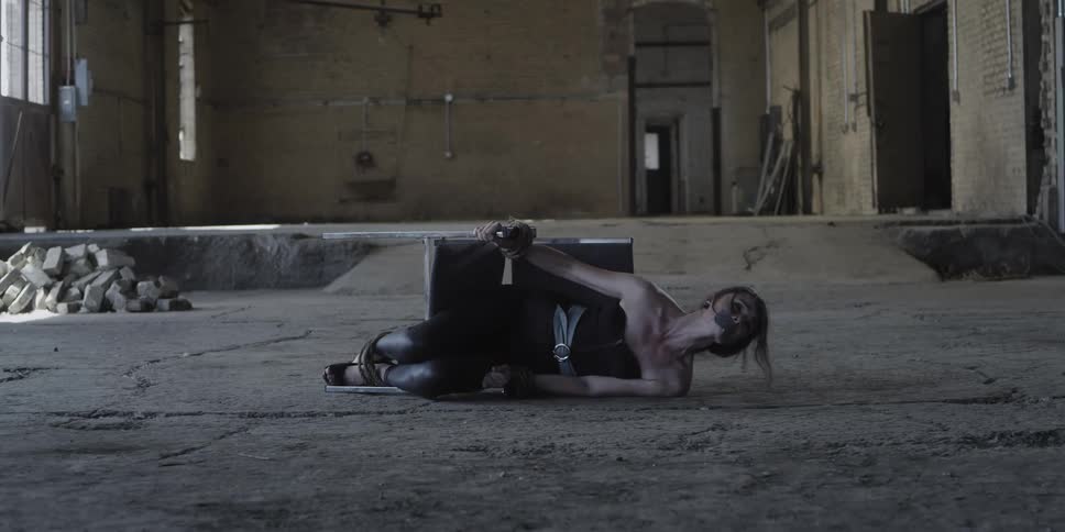 A woman in a room tied to a chair on the floor and mouth duct taped with a wall and doorway in the background