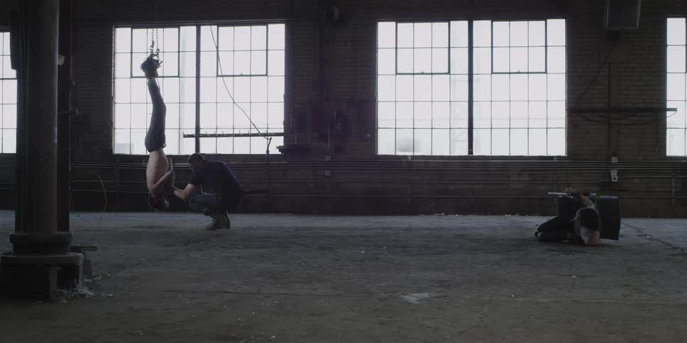 A man crouched on the floor messes with a man hanging by his feet while another person is tipped over in a chair on the floor