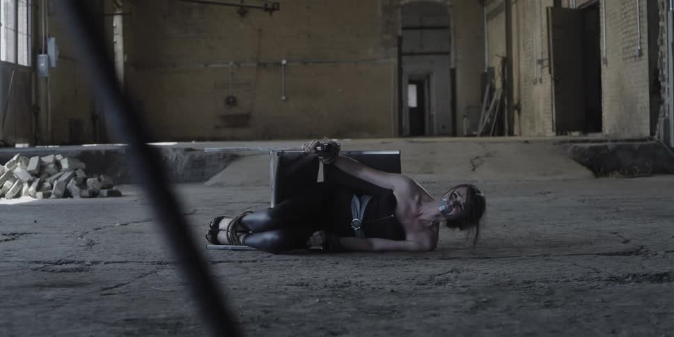 A woman in a room tied to a chair on the floor and mouth duct taped with a person with an ax in the foreground
