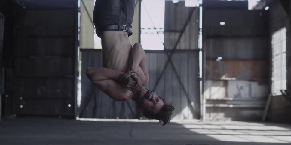 A shirtless man bound with rope and gagged is struggling to get free while hanging upside down in a warehouse during the day
