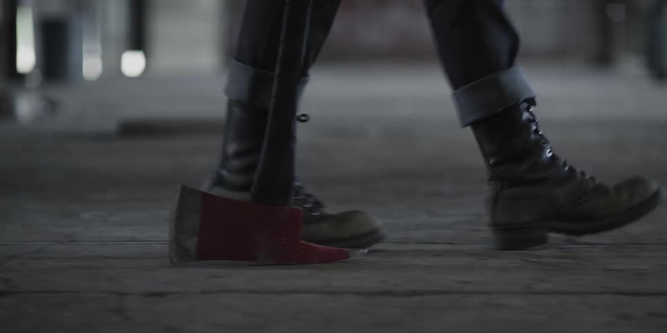 A person with boots walking with an ax in a room with a wall in the background