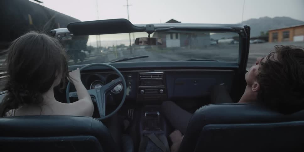A woman is driving a convertible with a man sleeping in the passenger seat as they travel alongside a train yard