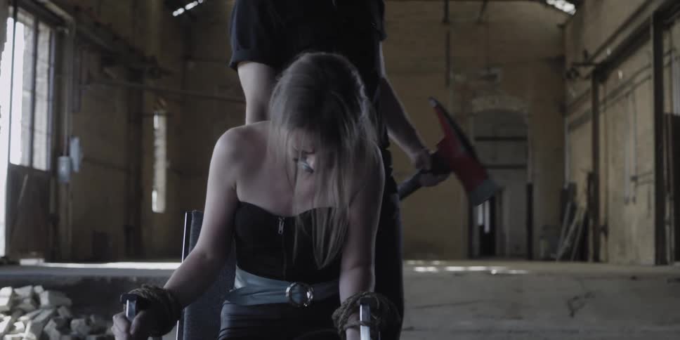 A man holding an axe drags a chair with a woman tied to it with her mouth gagged