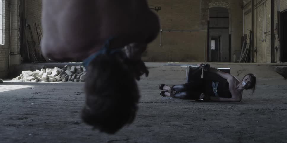 A man tied up upside down and a woman tied to a chair on the ground in a room with a wall in the background