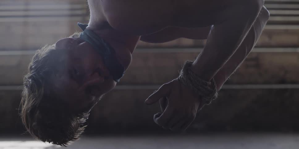 Shirtless scared gagged man tied up hanging upside down inside a warehouse