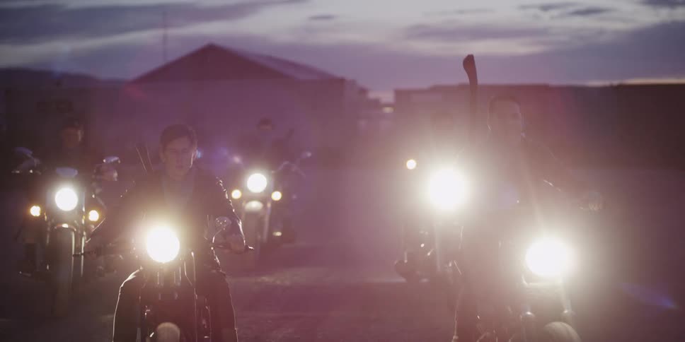 Men riding motorcycles with headlights with buildings the sky and clouds in the background