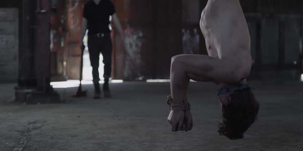 A man in a room tied up upside down as the man with an ax walk towards him with a wall and doorway in the background