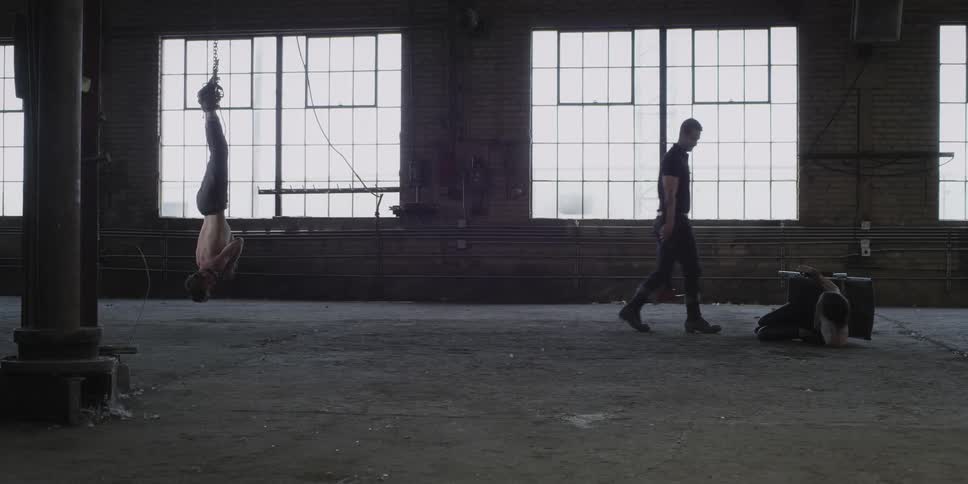 A man carrying an ax walks menacingly past two people who are bound and fearful