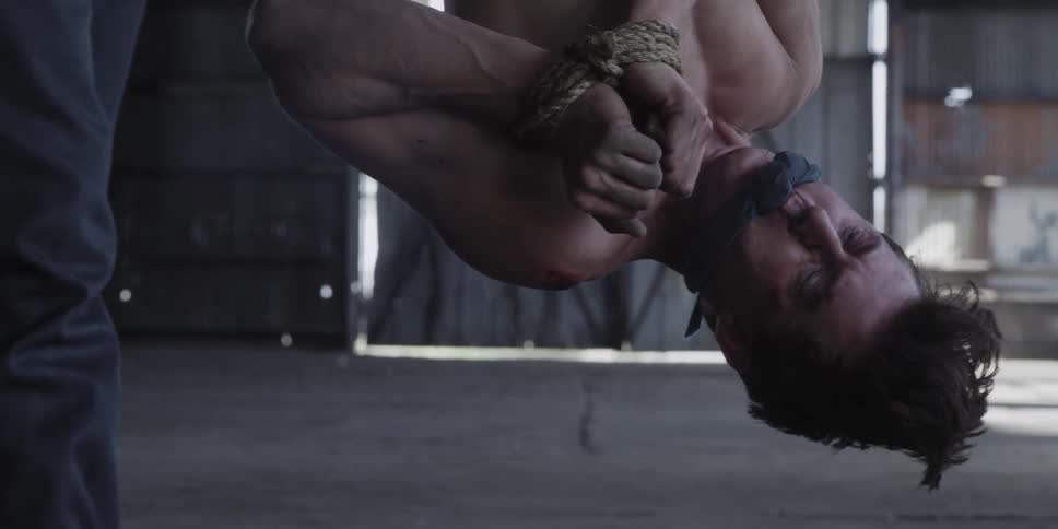 A man hanging upside down with his hands tied with rope as another man with an ax stoops down next to him