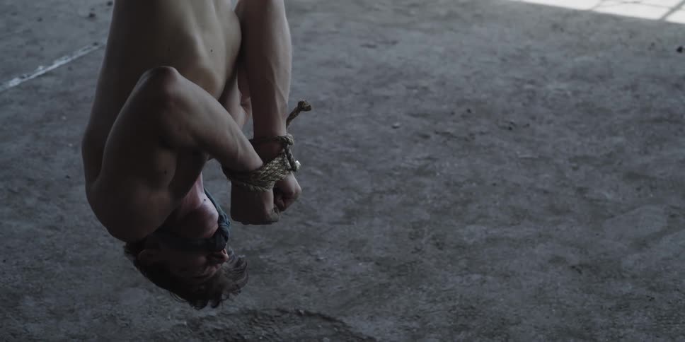 A man with his hands tied with rope hanging upside down with another man with an ax