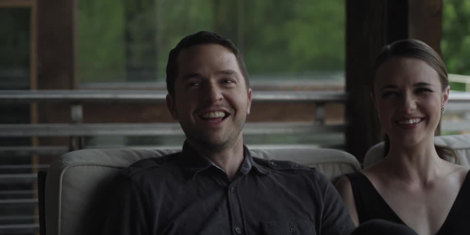 A man and woman in a room sitting on a couch with a wall and window in the background