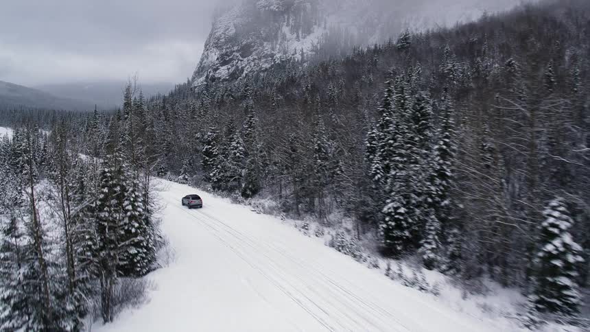 A silver automobile travels on a snow covered road surrounded by trees