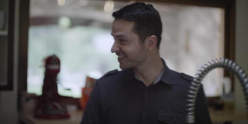 A man in a home kitchen is standing at a sink while having a pleasant conversation with someone else