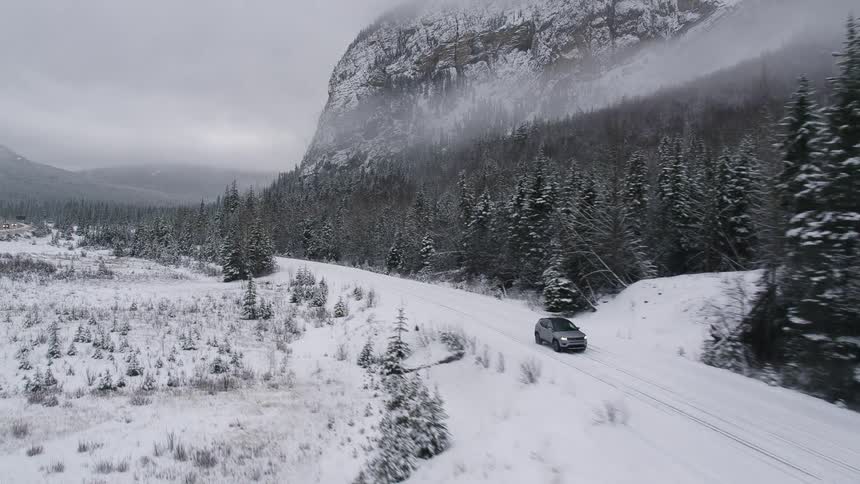A silver van travels on a snow covered road lined with trees and a mountain